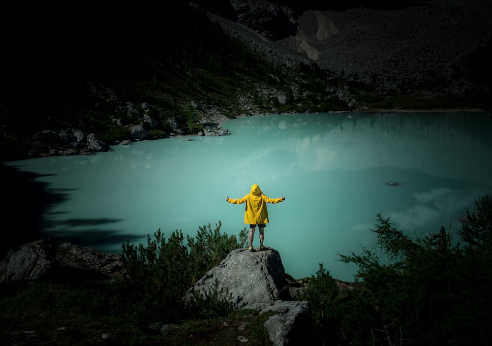 uomo in giacca gialla e jeans blu denim in piedi sulla roccia vicino allo specchio d'acqua durante