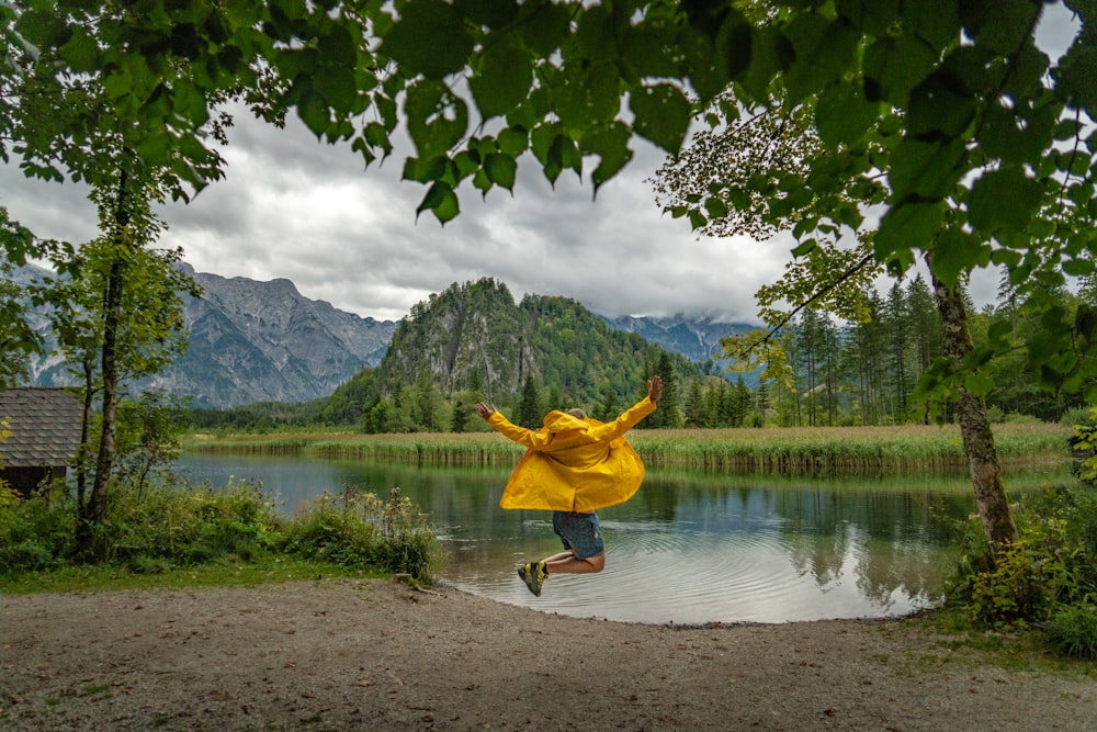 man in yellow jacket and blue denim jeans jumping on water near green trees and mountain