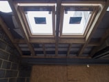 The image depicts the interior ceiling of a building under construction. There are wooden beams and a partial view of the roof structure. Two rectangular window installations are visible.