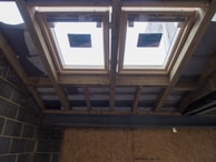 The image depicts the interior ceiling of a building under construction. There are wooden beams and a partial view of the roof structure. Two rectangular window installations are visible.