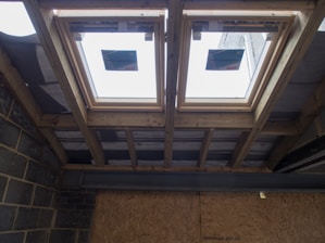 The image depicts the interior ceiling of a building under construction. There are wooden beams and a partial view of the roof structure. Two rectangular window installations are visible.