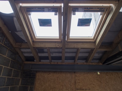 The image depicts the interior ceiling of a building under construction. There are wooden beams and a partial view of the roof structure. Two rectangular window installations are visible.