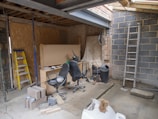 An indoor space appears to be under construction or renovation. Various items such as chairs, construction materials, and tools are scattered around. A wooden ladder leans against one wall, and a metal ladder stands nearby. The walls are partially finished with some areas revealing brickwork. A piece of large wood or drywall leans against the wall, and there is a black trash can nearby. The ceiling is exposed, showing wooden beams and some electrical wiring.