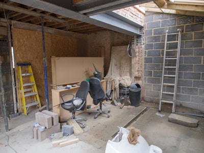 An indoor space appears to be under construction or renovation. Various items such as chairs, construction materials, and tools are scattered around. A wooden ladder leans against one wall, and a metal ladder stands nearby. The walls are partially finished with some areas revealing brickwork. A piece of large wood or drywall leans against the wall, and there is a black trash can nearby. The ceiling is exposed, showing wooden beams and some electrical wiring.