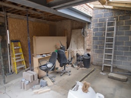 An indoor space appears to be under construction or renovation. Various items such as chairs, construction materials, and tools are scattered around. A wooden ladder leans against one wall, and a metal ladder stands nearby. The walls are partially finished with some areas revealing brickwork. A piece of large wood or drywall leans against the wall, and there is a black trash can nearby. The ceiling is exposed, showing wooden beams and some electrical wiring.