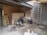 An indoor space appears to be under construction or renovation. Various items such as chairs, construction materials, and tools are scattered around. A wooden ladder leans against one wall, and a metal ladder stands nearby. The walls are partially finished with some areas revealing brickwork. A piece of large wood or drywall leans against the wall, and there is a black trash can nearby. The ceiling is exposed, showing wooden beams and some electrical wiring.