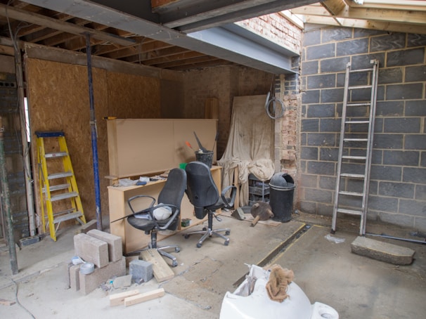 An indoor space appears to be under construction or renovation. Various items such as chairs, construction materials, and tools are scattered around. A wooden ladder leans against one wall, and a metal ladder stands nearby. The walls are partially finished with some areas revealing brickwork. A piece of large wood or drywall leans against the wall, and there is a black trash can nearby. The ceiling is exposed, showing wooden beams and some electrical wiring.