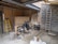 An indoor space appears to be under construction or renovation. Various items such as chairs, construction materials, and tools are scattered around. A wooden ladder leans against one wall, and a metal ladder stands nearby. The walls are partially finished with some areas revealing brickwork. A piece of large wood or drywall leans against the wall, and there is a black trash can nearby. The ceiling is exposed, showing wooden beams and some electrical wiring.