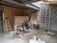 An indoor space appears to be under construction or renovation. Various items such as chairs, construction materials, and tools are scattered around. A wooden ladder leans against one wall, and a metal ladder stands nearby. The walls are partially finished with some areas revealing brickwork. A piece of large wood or drywall leans against the wall, and there is a black trash can nearby. The ceiling is exposed, showing wooden beams and some electrical wiring.