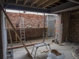 An unfinished construction site with exposed brick walls and scattered construction materials. A metal ladder leans against a wall, and a horizontal beam supports the ceiling. There are planks of wood, a metal pipe, and building blocks on the concrete floor. Tools and cables are visible around the site.