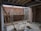An unfinished construction site with exposed brick walls and scattered construction materials. A metal ladder leans against a wall, and a horizontal beam supports the ceiling. There are planks of wood, a metal pipe, and building blocks on the concrete floor. Tools and cables are visible around the site.