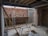An unfinished construction site with exposed brick walls and scattered construction materials. A metal ladder leans against a wall, and a horizontal beam supports the ceiling. There are planks of wood, a metal pipe, and building blocks on the concrete floor. Tools and cables are visible around the site.