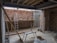 An unfinished construction site with exposed brick walls and scattered construction materials. A metal ladder leans against a wall, and a horizontal beam supports the ceiling. There are planks of wood, a metal pipe, and building blocks on the concrete floor. Tools and cables are visible around the site.