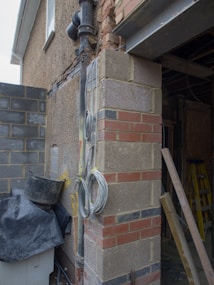 A construction site with exposed brickwork and utility wires coiled against a building wall. Various building materials such as bricks and stone blocks are present, along with a black tarp covering some items. A ladder is visible inside the structure.