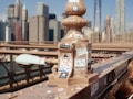 A decorative post on a bridge is adorned with various stickers, each displaying unique designs and messages. In the background, a city skyline with numerous skyscrapers stands tall under a clear blue sky. The image features the intricate metalwork of the bridge and highlights the urban scenery.