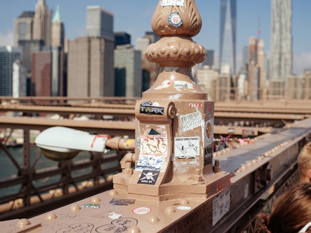 A decorative post on a bridge is adorned with various stickers, each displaying unique designs and messages. In the background, a city skyline with numerous skyscrapers stands tall under a clear blue sky. The image features the intricate metalwork of the bridge and highlights the urban scenery.