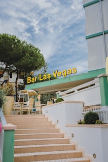 Outdoor seating area of a bar with clean stone steps leading up to a terrace. The building is modern with green and white accents. A large sign reading 'Bar Las Vegas' is prominently displayed. There are several tables and wicker chairs, with some greenery such as small bushes and trees, under a partly cloudy sky.