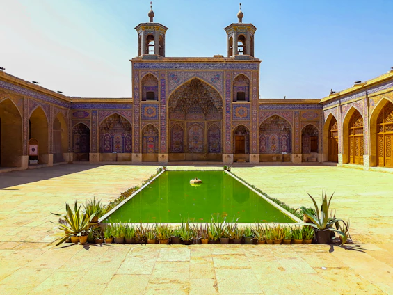 A warm Moroccan riad courtyard with zellige tiles, lush palm trees, and a sparkling pool reflecting the golden sunlight.