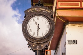 An old-fashioned clock with Roman numerals is mounted on a decorative, antique metal frame. The clock is attached to a white building, partially visible in the background, featuring a street sign that reads 'CALLE 5 CARRERA 6'. The sky is partially cloudy, and there is a part of a wooden structure with red and yellow trim visible on the right side.