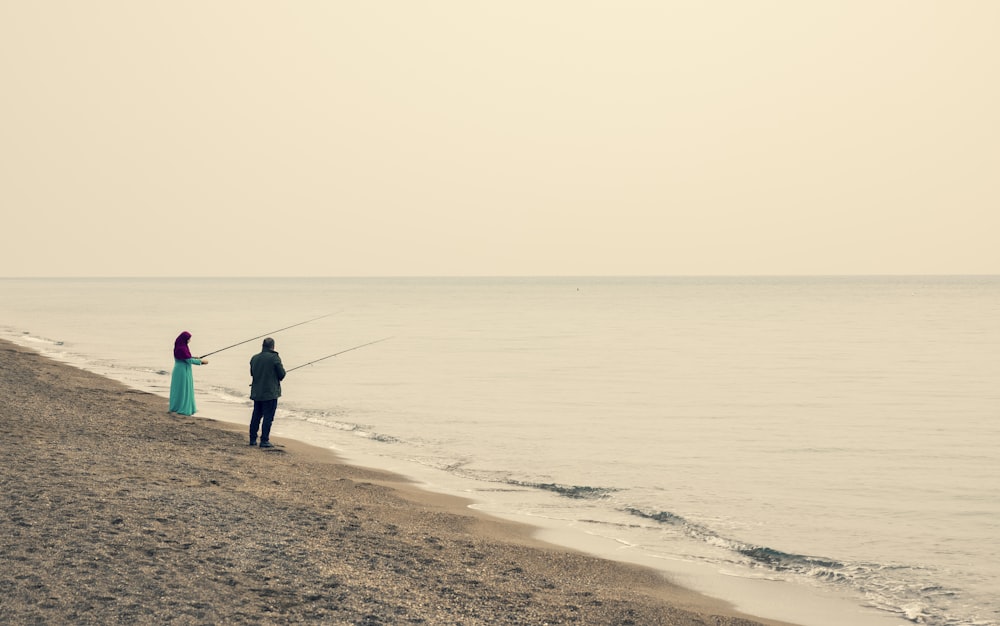 man in blue jacket and black pants holding fishing rod standing on seashore during daytime