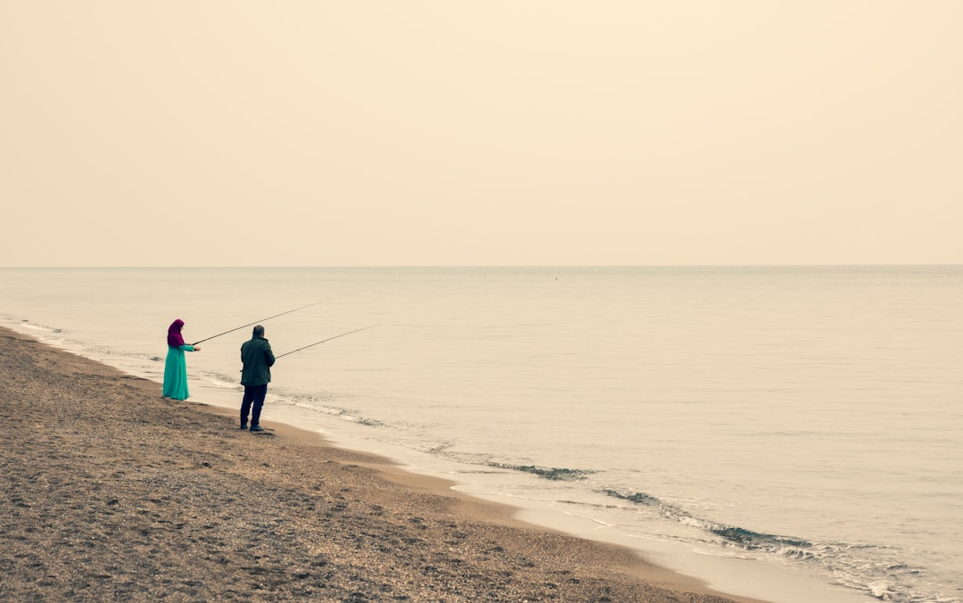 man in blue jacket and black pants holding fishing rod standing on seashore during daytime