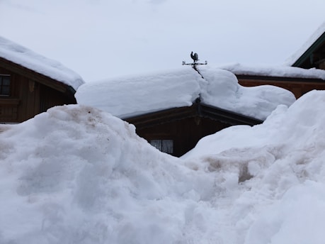 A snow-covered rooftop with a weather vane shaped like a rooster sits atop wooden buildings. Thick layers of snow blanket the structures and surrounding area, creating a wintery scene.