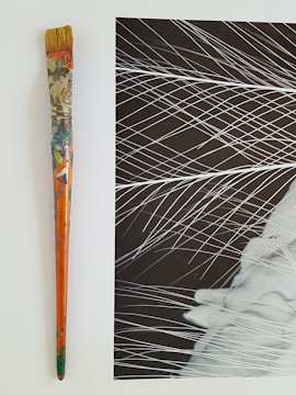 An aged paintbrush with a colorful, paint-splattered handle sits beside an abstract black and white artwork, featuring dynamic white lines crossing over a dark background.