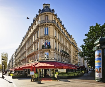 A classic Parisian building with ornate architecture is situated on a street corner. It features several stories, a mansard roof, and decorative railings. The ground floor is occupied by a restaurant with red awnings and an outdoor seating area. A lamppost and a cylindrical advertising column are visible along the street. The scene is illuminated by bright sunlight, and a bird is flying in the sky.
