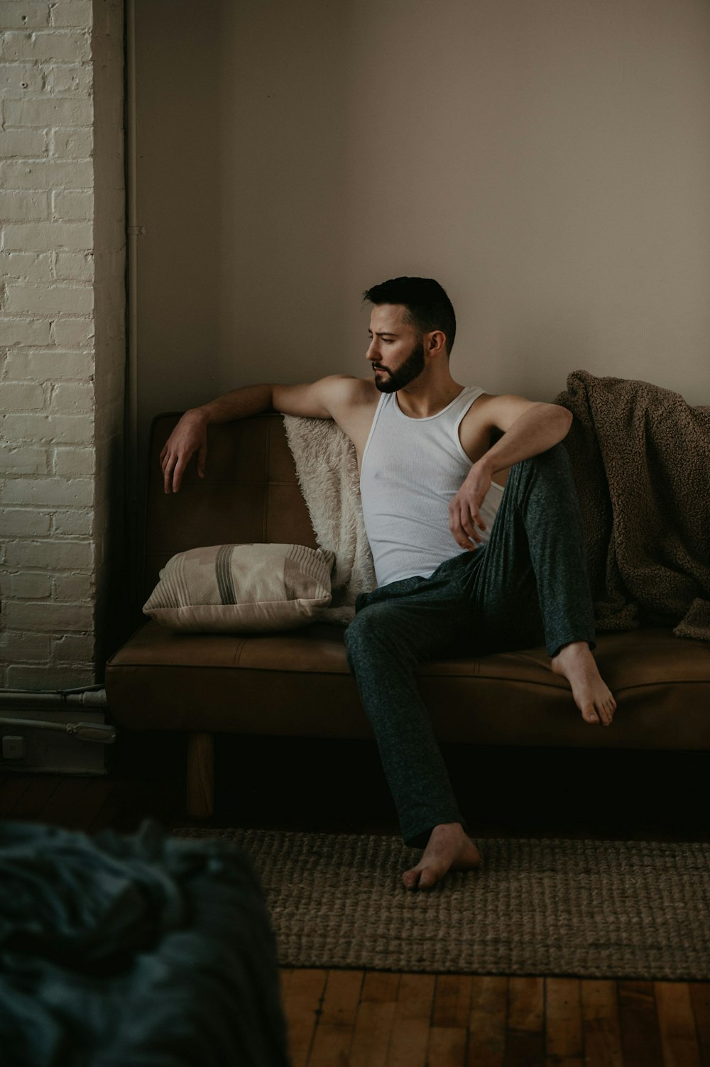 man in white tank top and blue denim jeans sitting on brown sofa
