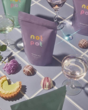 Several colorful packages labeled 'not pot' are arranged on a tiled surface alongside an assortment of gourmet chocolates, a tart with green filling, delicate purple flowers, and elegant glassware containing light beverages. The soft lighting enhances the pastel hues, creating a dreamy, inviting atmosphere.