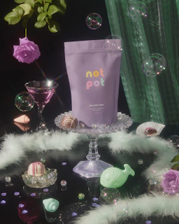 A dreamy flat lay of magic roll mushroom prerolls surrounded by fresh botanicals like blue lotus and herbs with a purple liquid swirl background