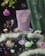 A whimsical and dreamy setup with a lavender-colored pouch labeled 'not pot' placed on a glass pedestal. Surrounding the pouch are various decorative elements including a martini glass with a purple liquid, a pink rose, chocolates, and a green fish-shaped object. Bubbles float around, adding to the enchanting atmosphere, while mint green feathers and small gem-like objects accompany the scene.