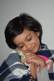 A young child with short black hair gently hugs a small stuffed animal with a content expression. The child is wrapped in a blue and white checkered blanket, and the background is a plain light color, creating a calm and cozy atmosphere.