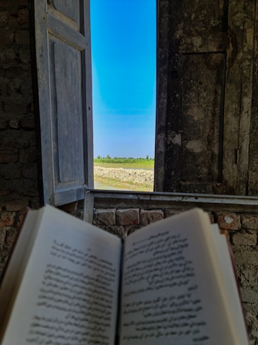 An open book with text in focus is held in front of a rustic wooden window. The window frames a clear blue sky and a green landscape with trees in the distance. The wall surrounding the window appears to be made of rough stone or brick.