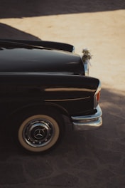 A classic black car is parked on a sunlit cobblestone surface. The car's body reflects the ambient light, creating a gleaming effect along the side. Decorative floral arrangements are attached to the front, suggesting a celebratory or ceremonial context. The vintage look of the car is accentuated by its chrome details and white-walled tires.