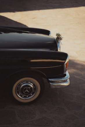 A classic black car is parked on a sunlit cobblestone surface. The car's body reflects the ambient light, creating a gleaming effect along the side. Decorative floral arrangements are attached to the front, suggesting a celebratory or ceremonial context. The vintage look of the car is accentuated by its chrome details and white-walled tires.