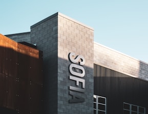 The image features a modern building with a mix of materials including concrete blocks and metal panels. The building displays the word 'SOFIA' in large, bold letters on its facade, accompanied by the word 'THE' above it. The structure is bathed in sunlight, creating a play of light and shadow across its surfaces.