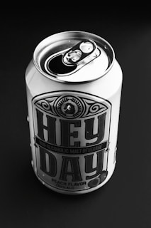 A can of non-alcoholic malt beverage labeled 'Hey Day' with peach flavor, placed on a dark background. The can is metal with a pop-top lid, prominently featuring bold typography and decorative design elements.