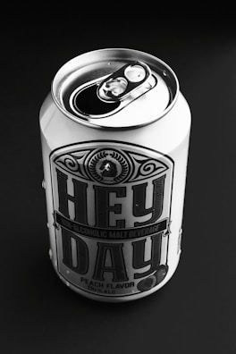 A can of non-alcoholic malt beverage labeled 'Hey Day' with peach flavor, placed on a dark background. The can is metal with a pop-top lid, prominently featuring bold typography and decorative design elements.