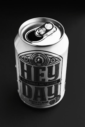 A can of non-alcoholic malt beverage labeled 'Hey Day' with peach flavor, placed on a dark background. The can is metal with a pop-top lid, prominently featuring bold typography and decorative design elements.