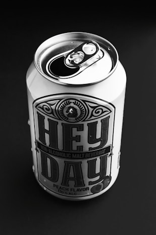 A can of non-alcoholic malt beverage labeled 'Hey Day' with peach flavor, placed on a dark background. The can is metal with a pop-top lid, prominently featuring bold typography and decorative design elements.