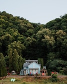 A picturesque blue house surrounded by lush green trees with a large forested hill in the background. The house has a charming appearance with red chairs on the porch and a gazebo nearby. There are two kayaks resting on the grass, adding a touch of outdoor recreation to the scene.