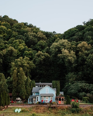 A picturesque blue house surrounded by lush green trees with a large forested hill in the background. The house has a charming appearance with red chairs on the porch and a gazebo nearby. There are two kayaks resting on the grass, adding a touch of outdoor recreation to the scene.