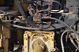 A close-up view of a hydraulic machine component with several interconnected metal pipes and hoses. The machinery appears heavily used, with visible dirt and oily residue coating parts of the structure. The main focus is on a central gear or joint caked in mud and oil, surrounded by a network of thick black cables and yellow metal framework.