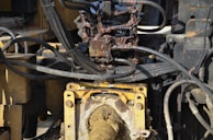 A close-up view of a hydraulic machine component with several interconnected metal pipes and hoses. The machinery appears heavily used, with visible dirt and oily residue coating parts of the structure. The main focus is on a central gear or joint caked in mud and oil, surrounded by a network of thick black cables and yellow metal framework.