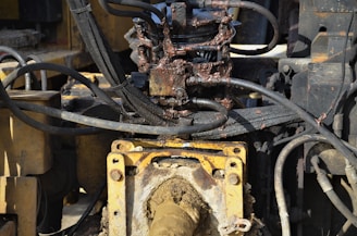 A close-up view of a hydraulic machine component with several interconnected metal pipes and hoses. The machinery appears heavily used, with visible dirt and oily residue coating parts of the structure. The main focus is on a central gear or joint caked in mud and oil, surrounded by a network of thick black cables and yellow metal framework.