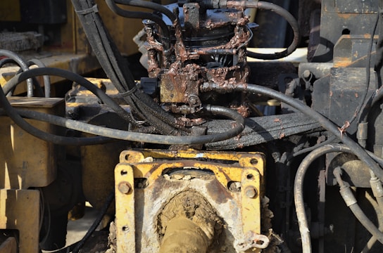 A close-up view of a hydraulic machine component with several interconnected metal pipes and hoses. The machinery appears heavily used, with visible dirt and oily residue coating parts of the structure. The main focus is on a central gear or joint caked in mud and oil, surrounded by a network of thick black cables and yellow metal framework.