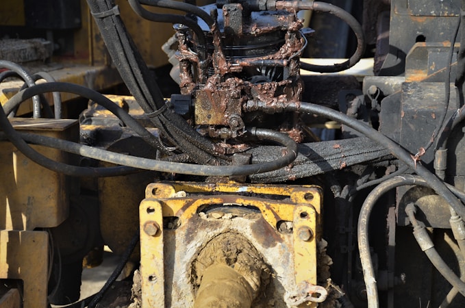 A close-up view of a hydraulic machine component with several interconnected metal pipes and hoses. The machinery appears heavily used, with visible dirt and oily residue coating parts of the structure. The main focus is on a central gear or joint caked in mud and oil, surrounded by a network of thick black cables and yellow metal framework.