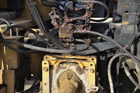 A close-up view of a hydraulic machine component with several interconnected metal pipes and hoses. The machinery appears heavily used, with visible dirt and oily residue coating parts of the structure. The main focus is on a central gear or joint caked in mud and oil, surrounded by a network of thick black cables and yellow metal framework.