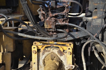 A close-up view of a hydraulic machine component with several interconnected metal pipes and hoses. The machinery appears heavily used, with visible dirt and oily residue coating parts of the structure. The main focus is on a central gear or joint caked in mud and oil, surrounded by a network of thick black cables and yellow metal framework.